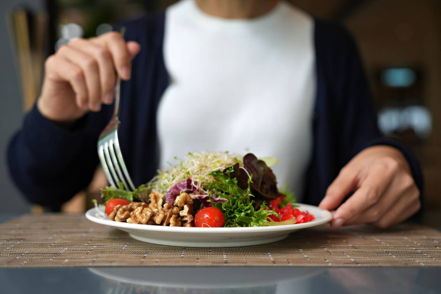A plate of vegan salad featuring healthy plant-based foods including nuts, greens, and tomatoes, and alfalfa sprouts, with the body of a woman visible sitting behind the plate